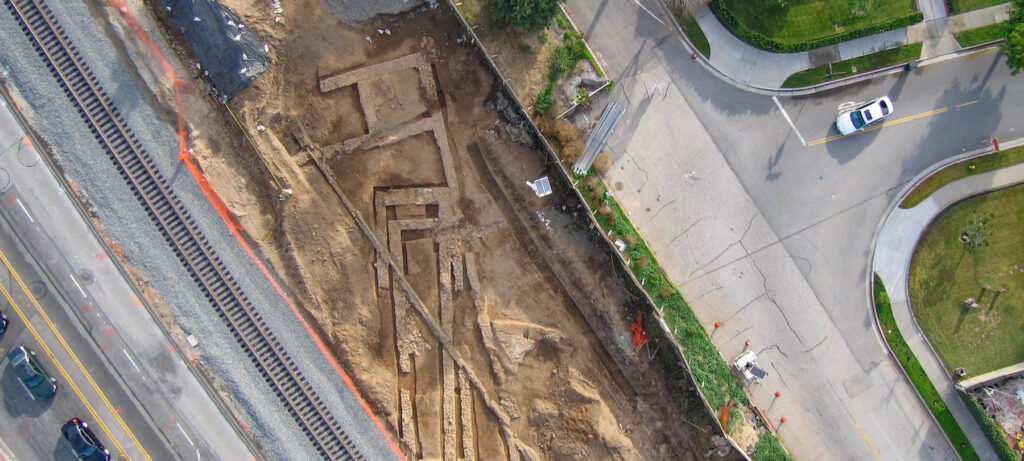 Aerial view of a long trench with exposed pipes, dug between a road, railroad, and a sidewalk.