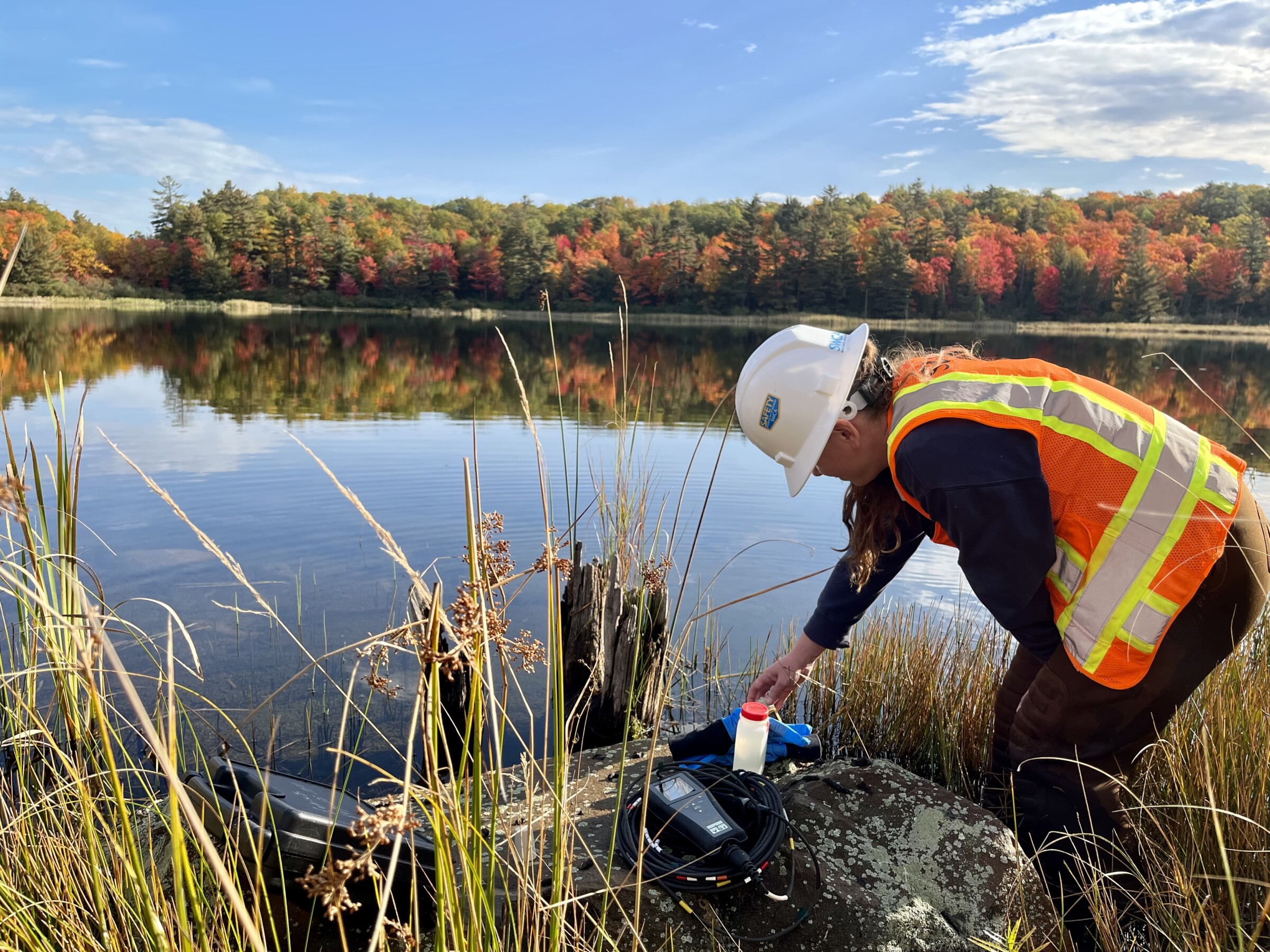A person wearing an orange safety vest and a hardhat sampling a body of water with colorful trees in the background.