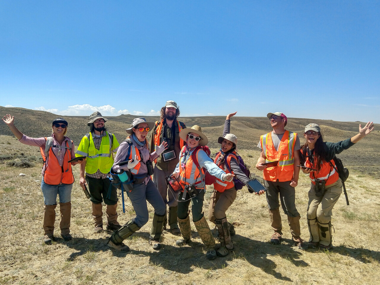A group of 8 people wearing safety vests and gear smiling and holding out their arms for a picture.