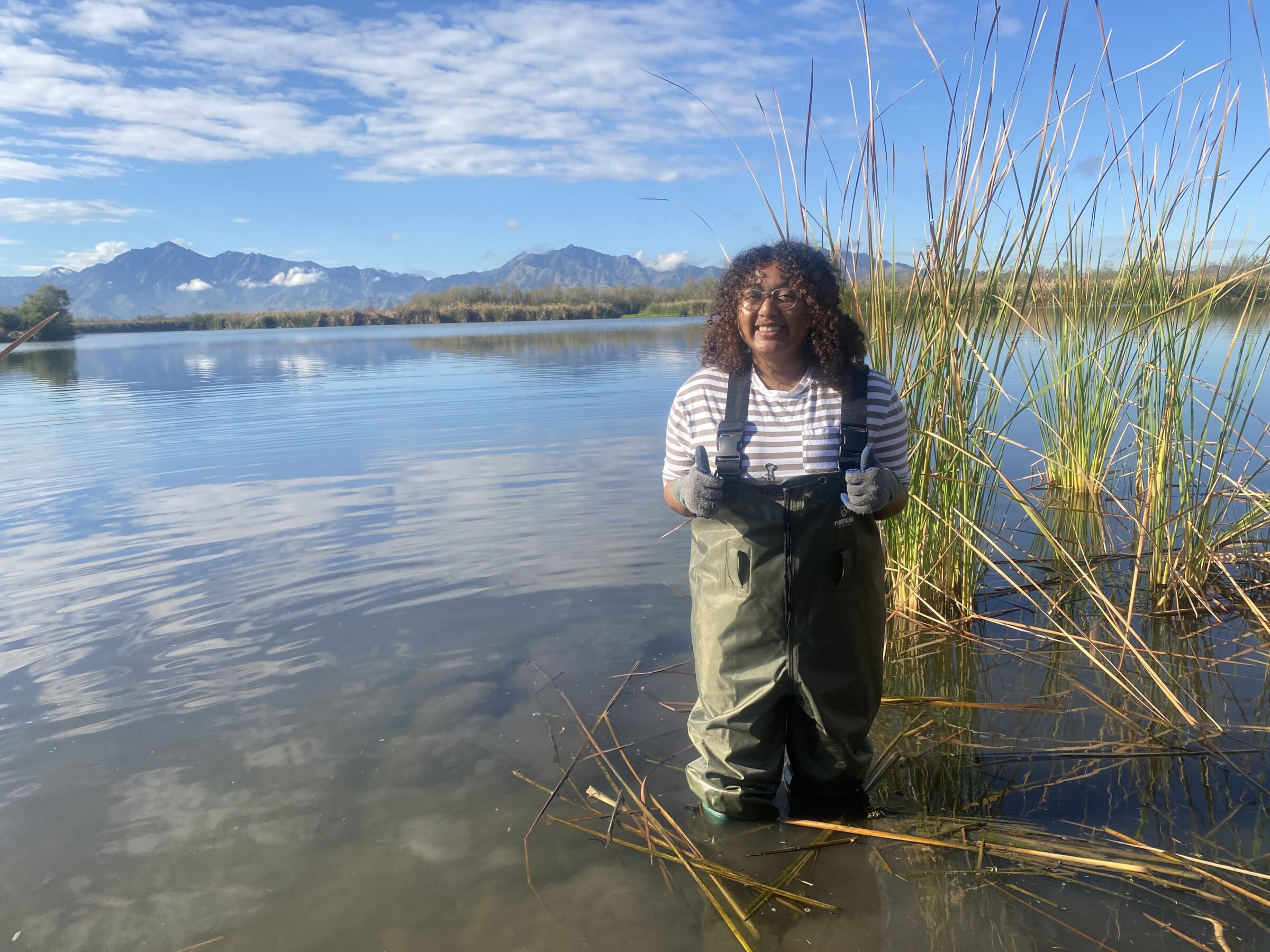 Raisa in a knee-deep body of water with vegetation and a mountain in the background. 
