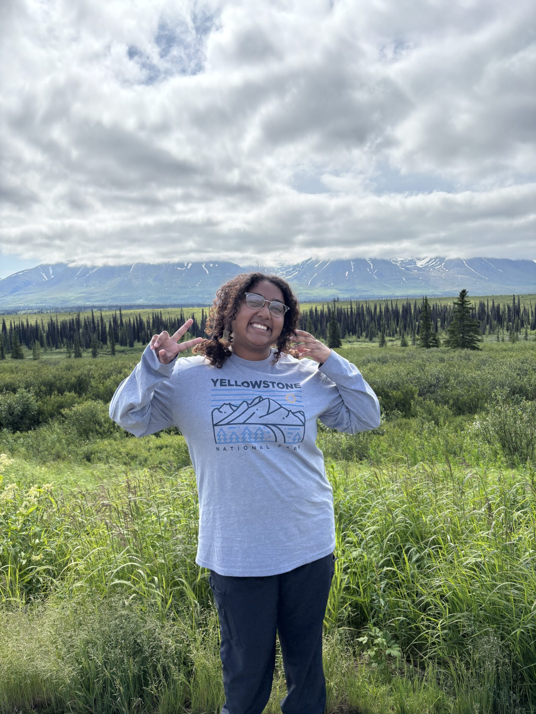 Raisa standing in vegetation with trees, a mountain, and clouds in the background. 