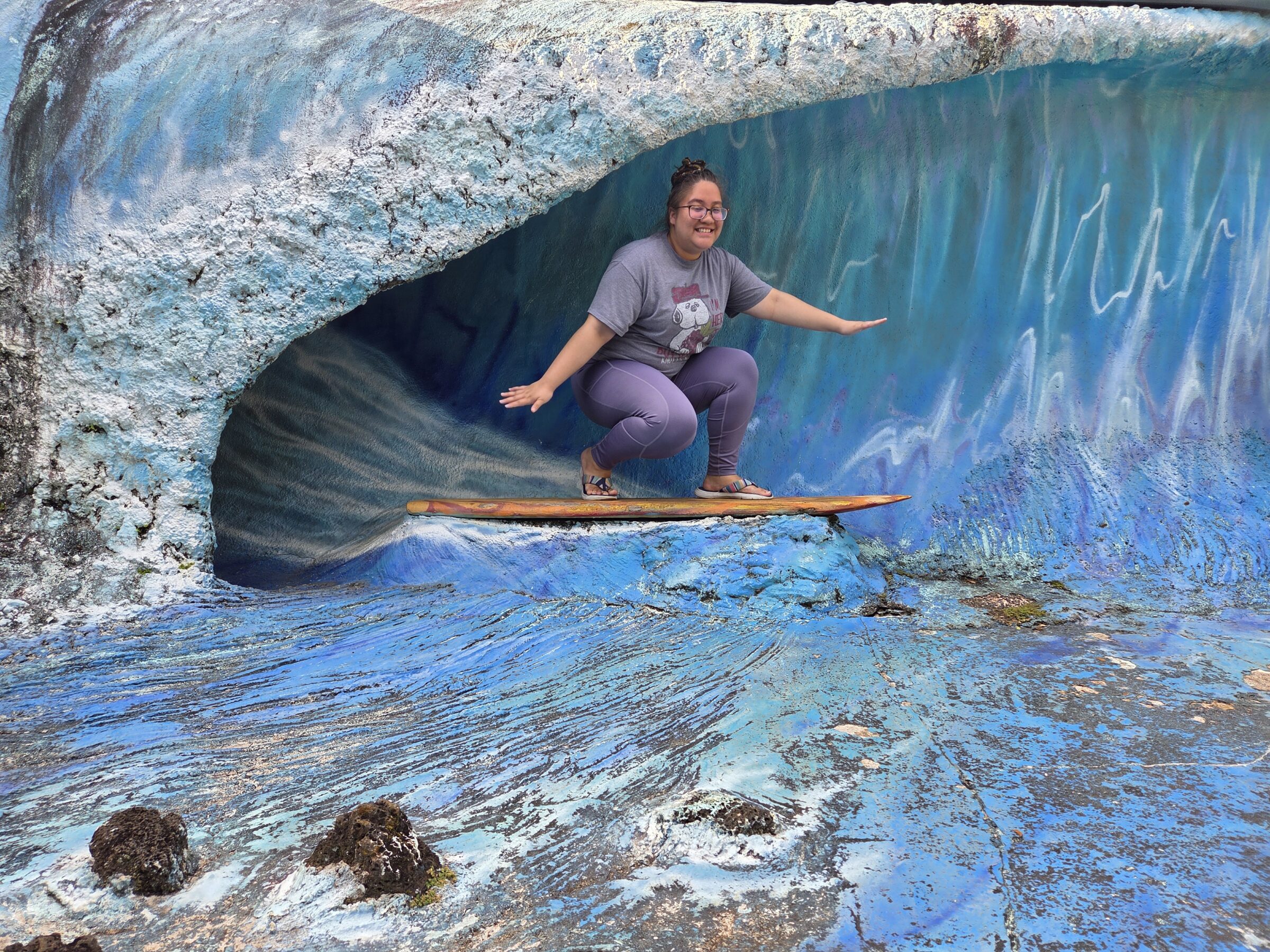 Shayla standing on a stationary surfboard on a tourist picture backdrop that looks like a blue wave in Kauaʻi. 