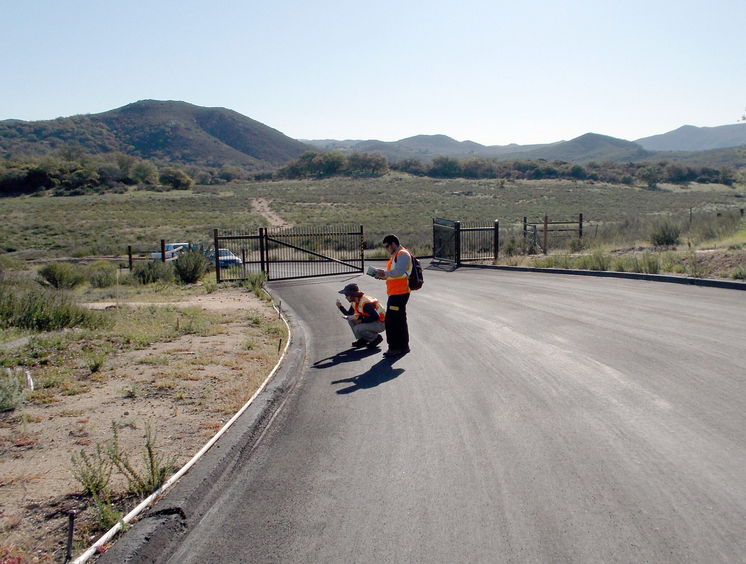Two people in orange safety vest on a paved road with a gate slightly open behind them, collecting field data.