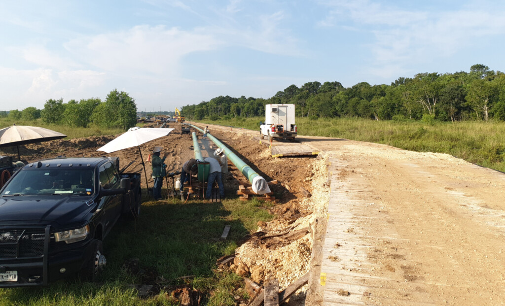 Contractors working on steel pipes with a truck and road next to them.