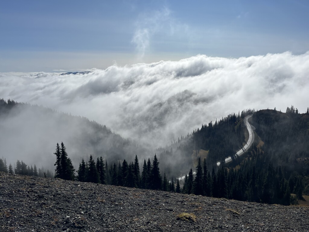 Mountains with a winding road on one of them, tress all around, and clouds all around.