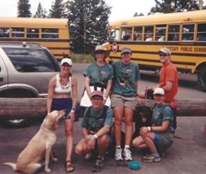 Team photo from early 2000s, featuring members of the Flagstaff office. Shows 6 people sitting on a wood log with a dog and school buses and cars behind them. 