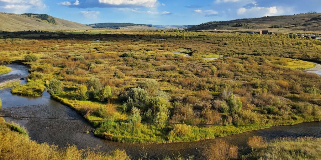 A wide view of a winding creek with vegetation growing all around it. Mountains are seen in the background.