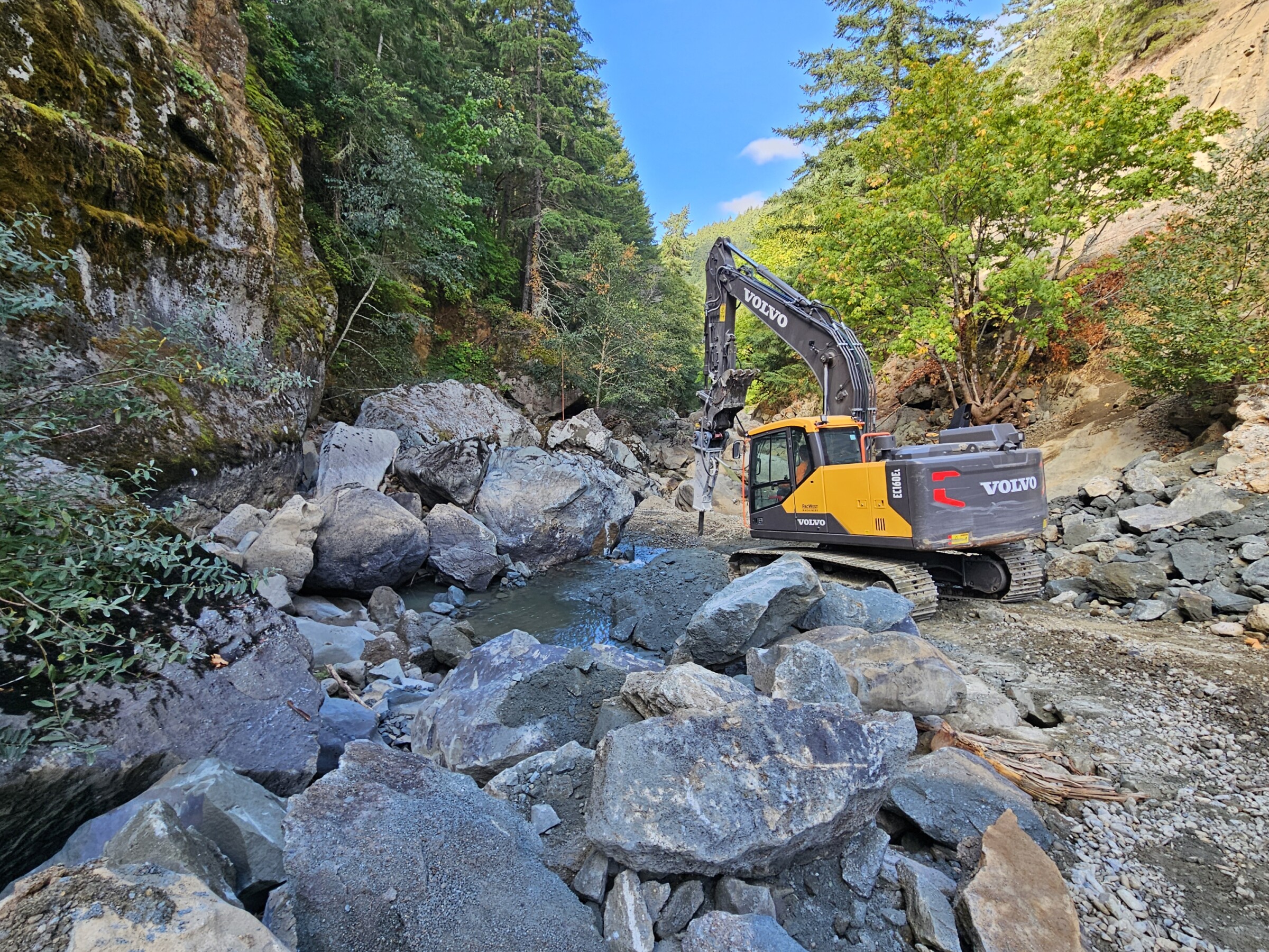 An excavator uses a pneumatic hammer to break boulders on the rivers path.