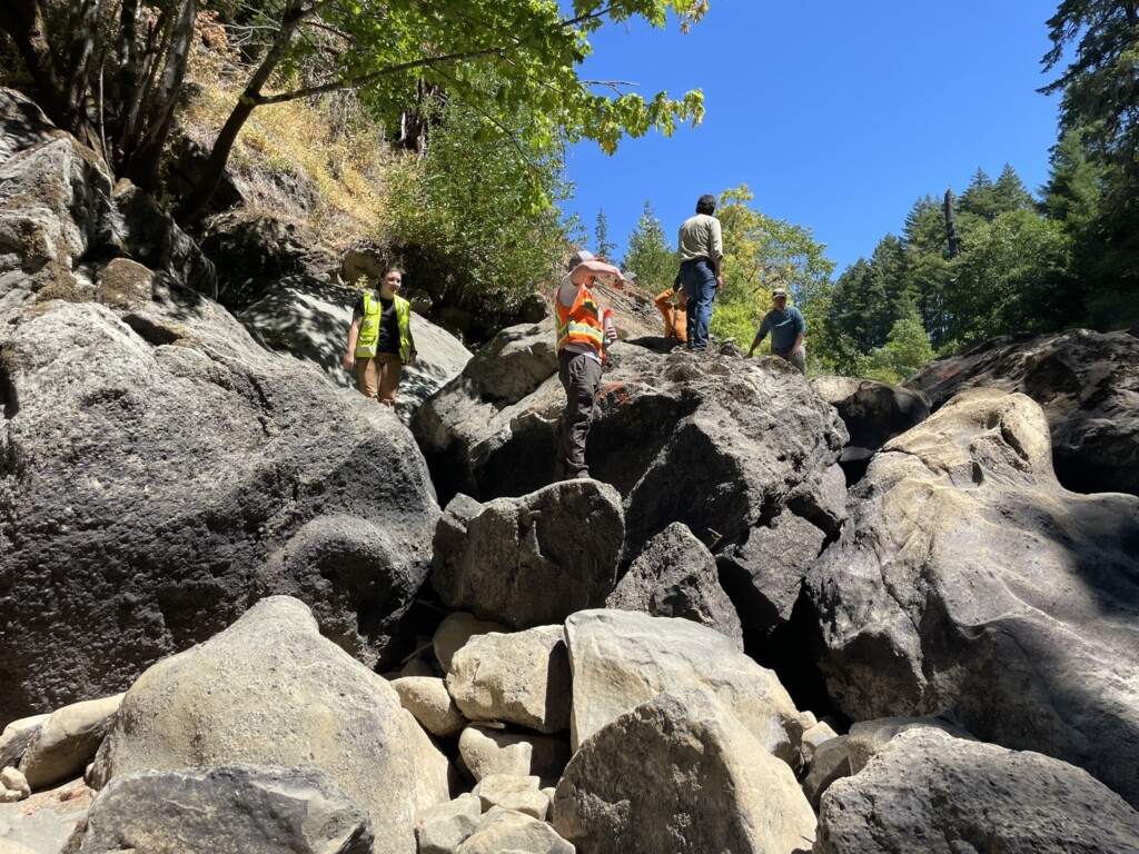 Several people accessing site conditions during a pre-construction meeting. They are all standing on large rocks and boulders wearing safety vests. 
