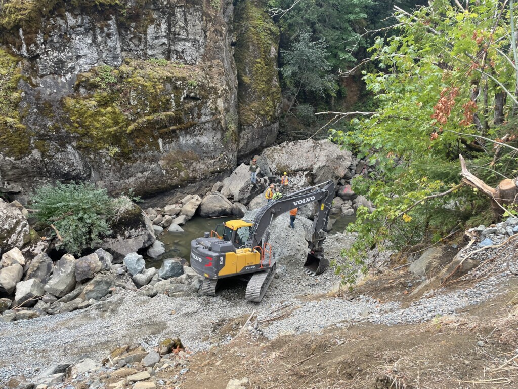 Heavy earthwork equipment (excavator) entering the middle fork coquille river canyon channel. Boulders are seen in the rivers. 
