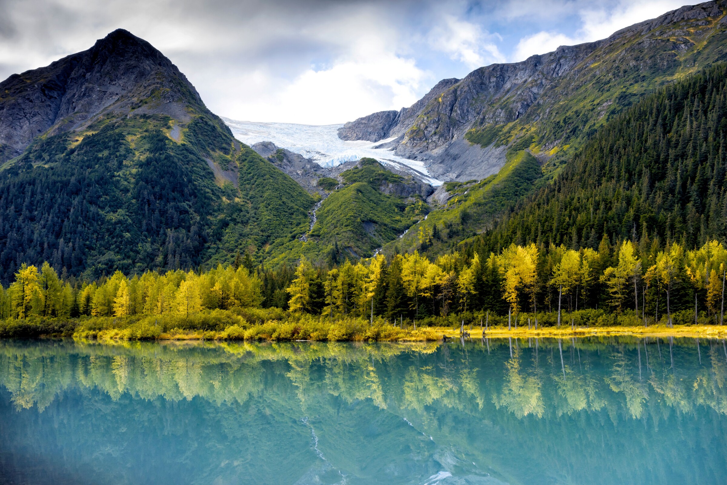 An image of the Anchorage Alaska state park showing wa body of water with green trees along the coast and a mountain with snow not far behind.