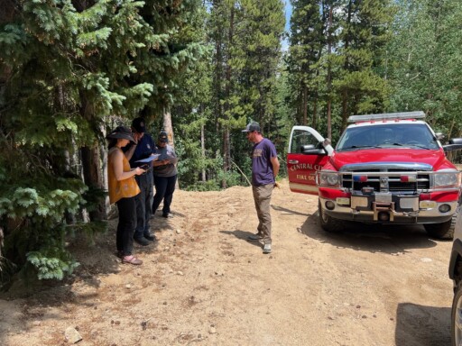 Four people standing next to a fire department red service vehicle and trees are surrounding them. They are on a dirt path taking notes and looking at their phones.