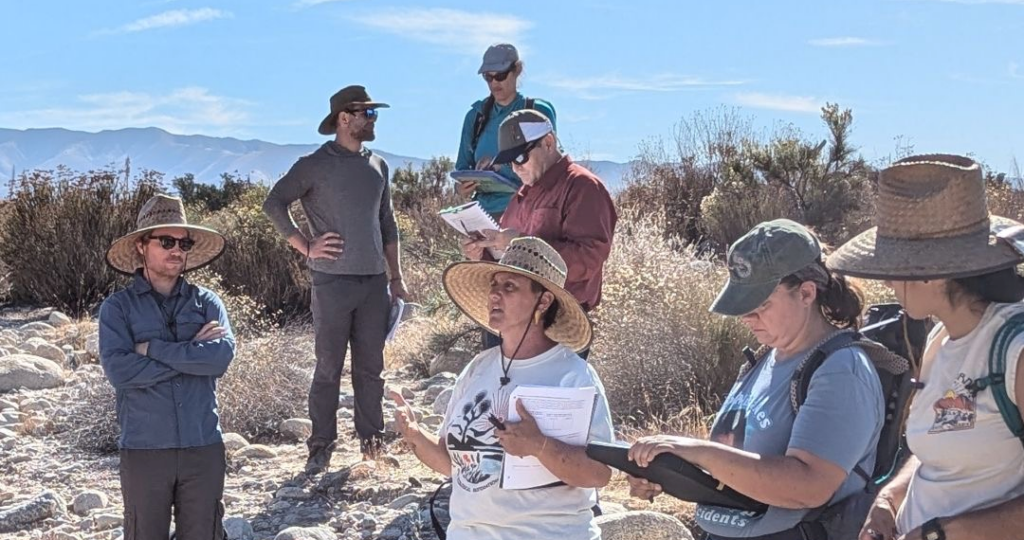 Several people standing outside with hands, glasses, and writing materials.