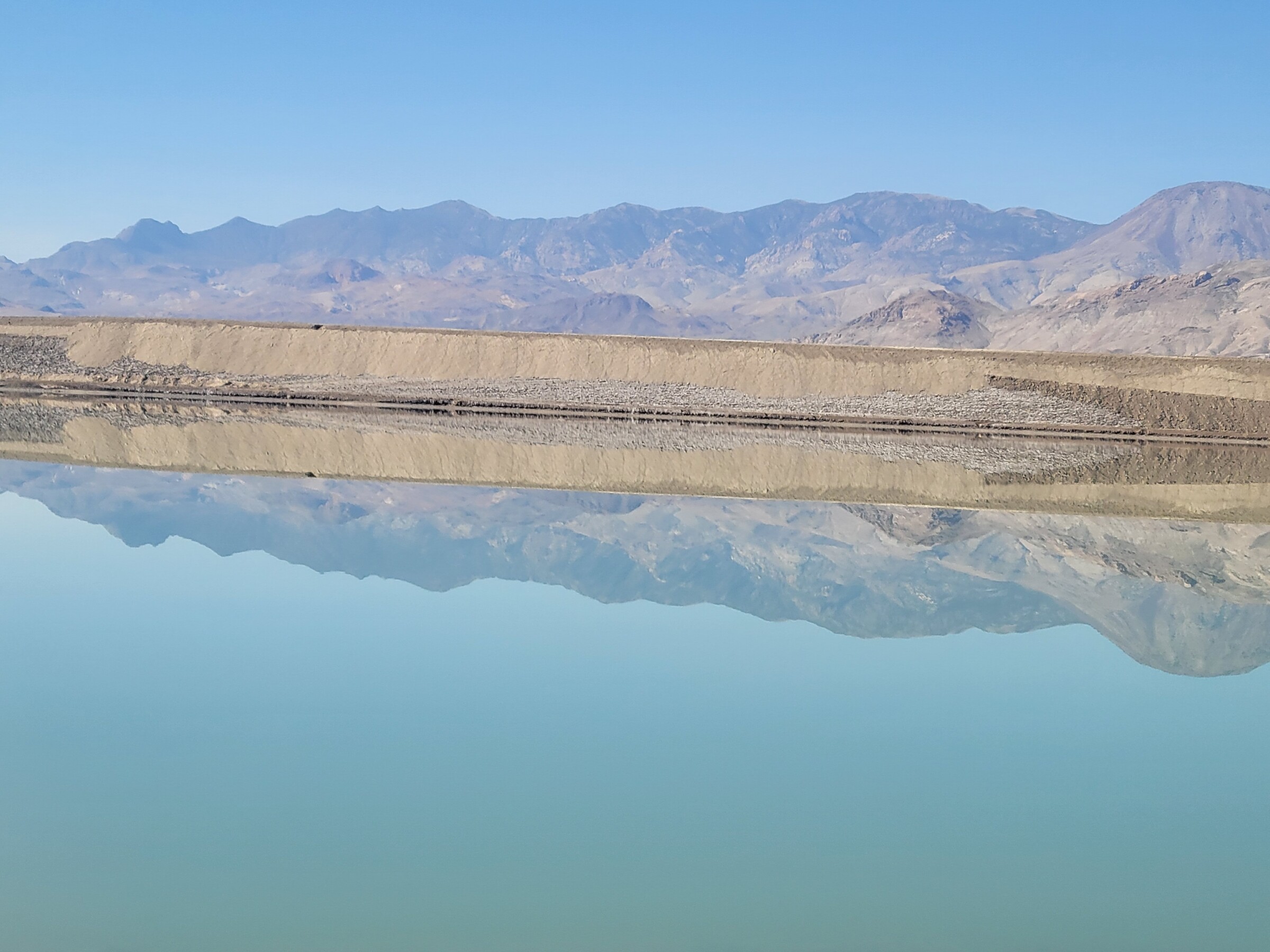 The sky reflects off a lithium evaporation pond in the foreground. A salt storage pile is visible in the background.
