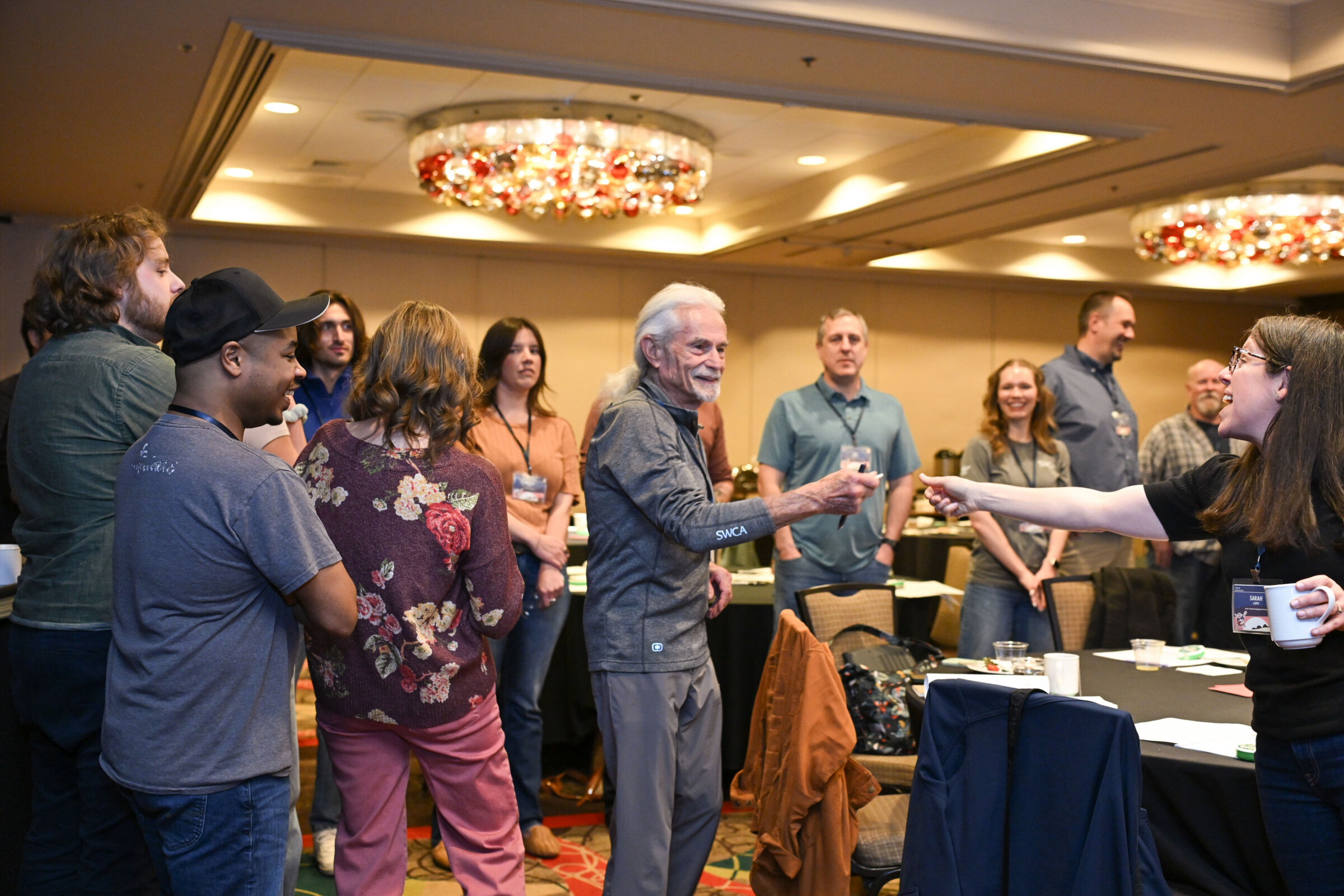 Several people standing in a lit-up conference room with table sand chair around them. Steven W. Carother's, Founder, can be seen handing Sara Lupis, Facilitator, a piece of paper.