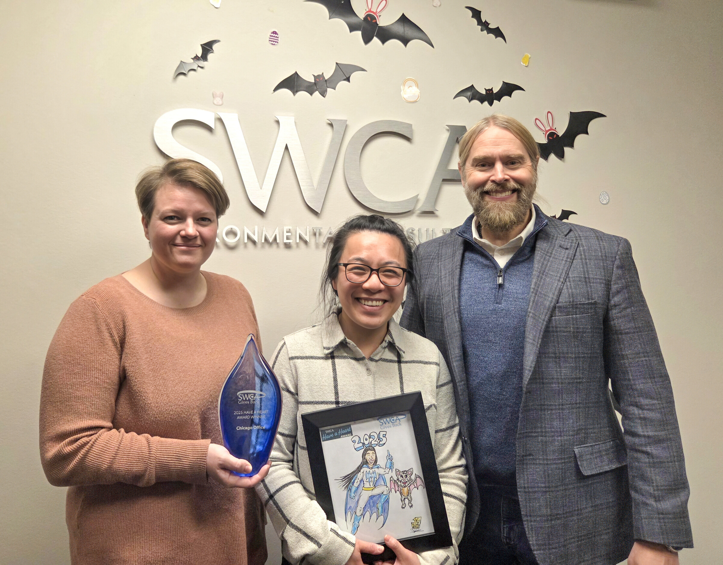 Three people standing in front of a SWCA Environmental consultant's sign. One person is Holding a blue transparent trophy and the other holds a framed drawing in their hand.