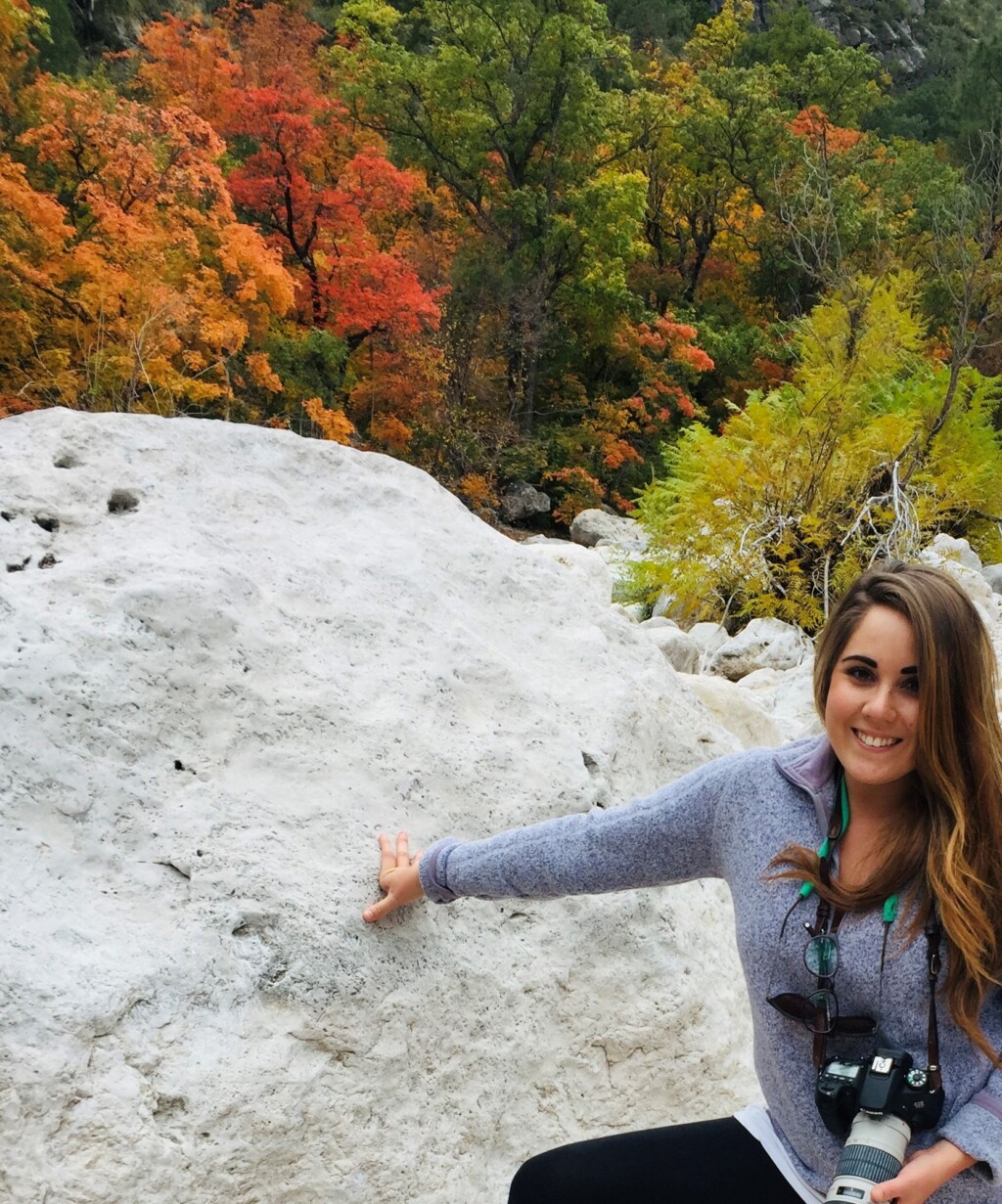 Sydney Lance with a camera standing on a large rock with colorful tress in the background. 
